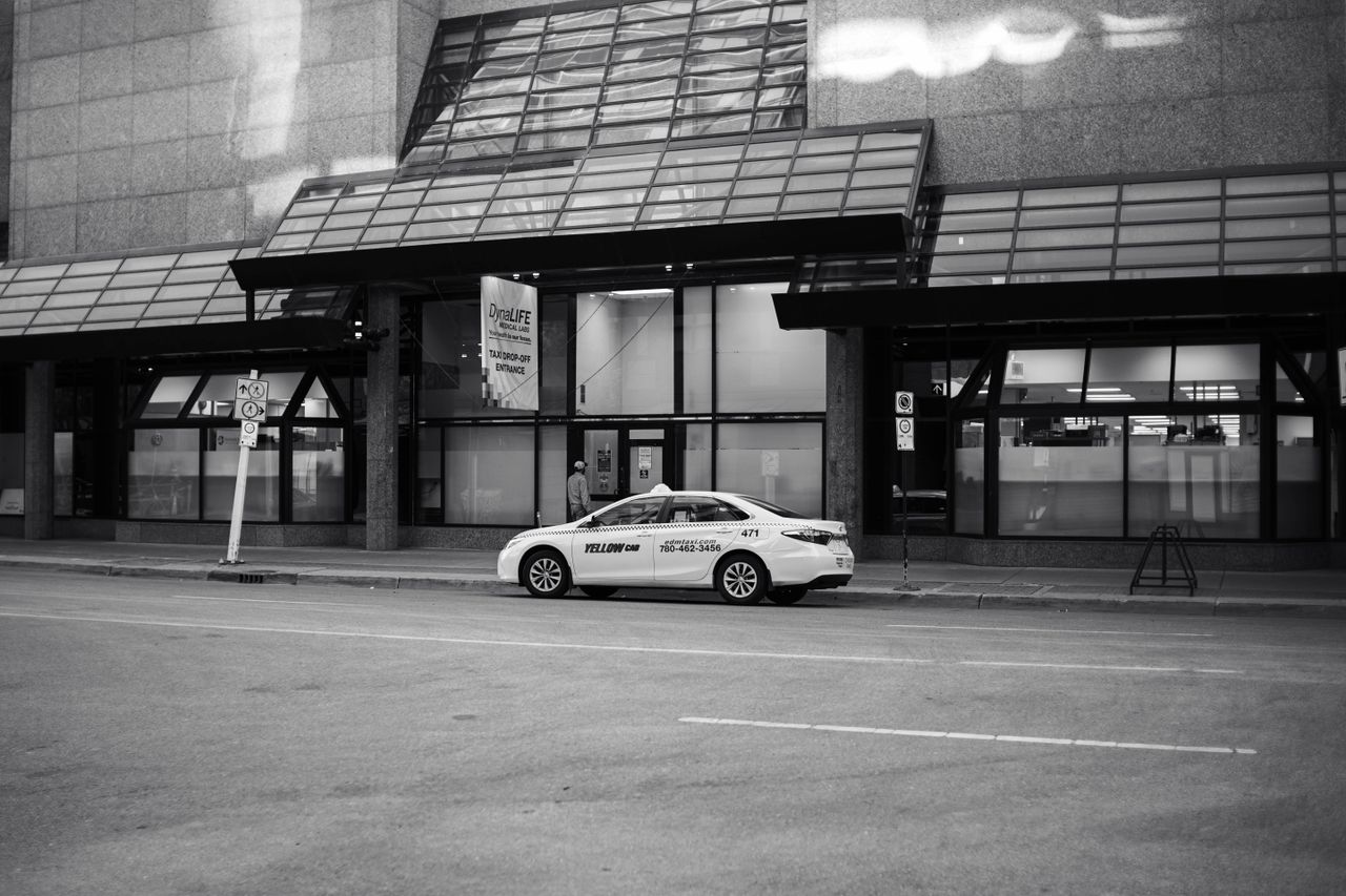 gallery-1 Black and white urban street scene featuring a parked taxi by modern glass buildings.
