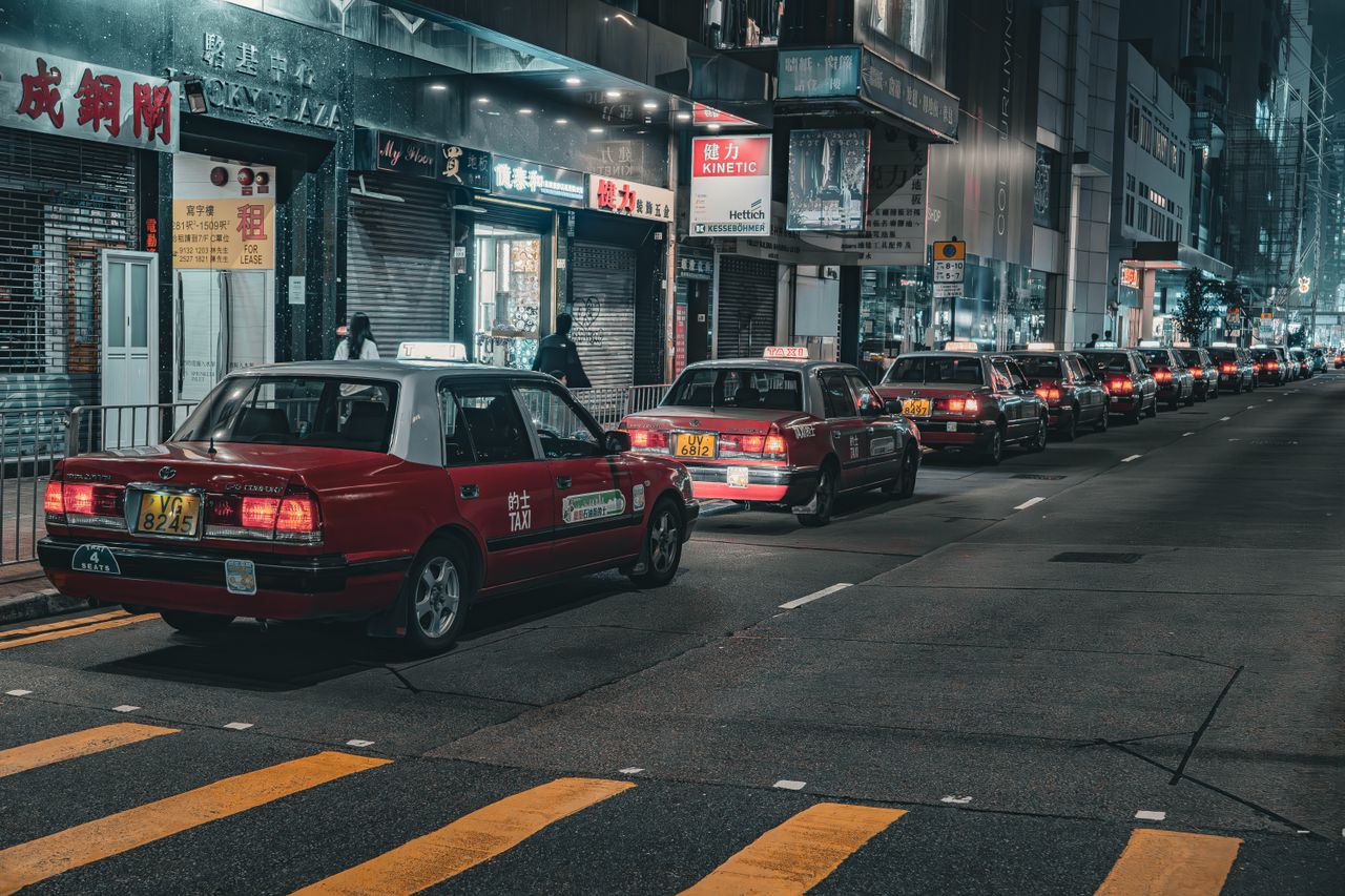 hero-img-02 A street in Hong Kong bustling with red taxis at night, capturing urban life and city lights.
